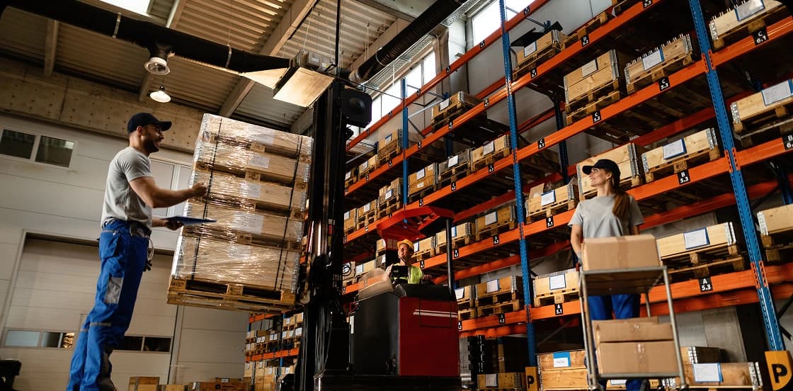 Warehouse workers operating forklift to move pallets on high shelving racks in large distribution center